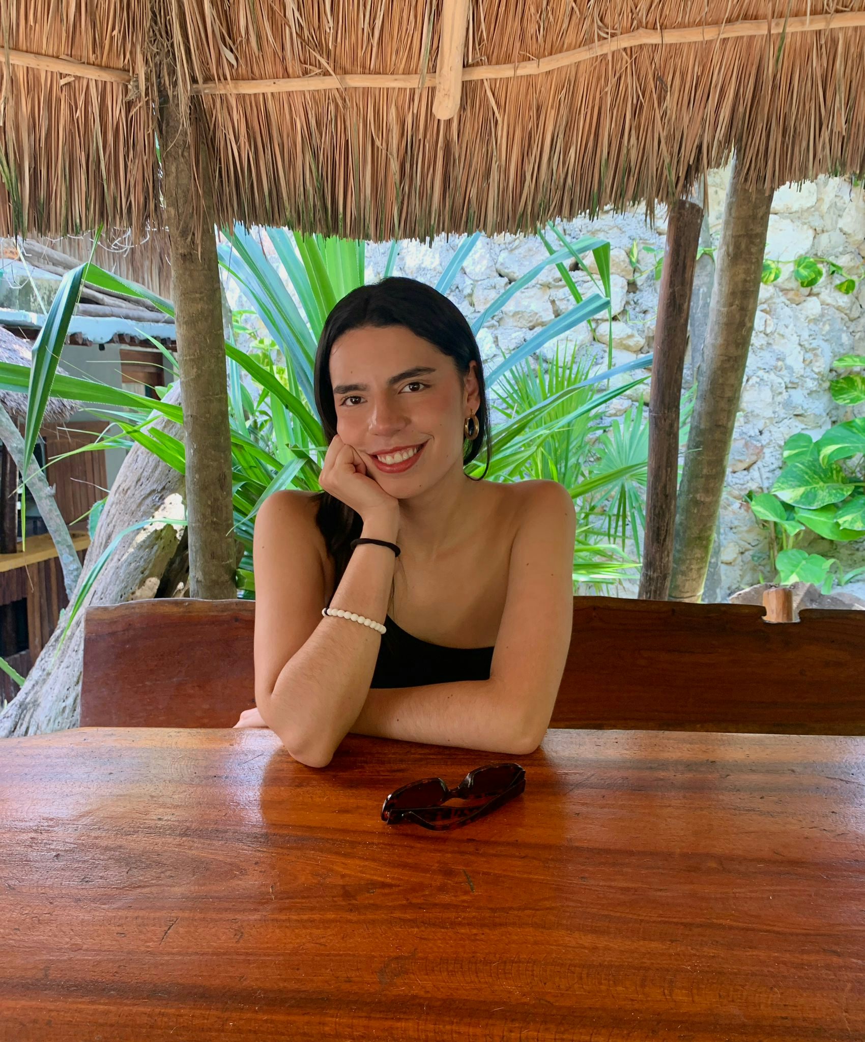 Travel Advisor Daniela Rodriguez sits with her head in her hand at a wood table under a straw umbrella with tropical trees behind her