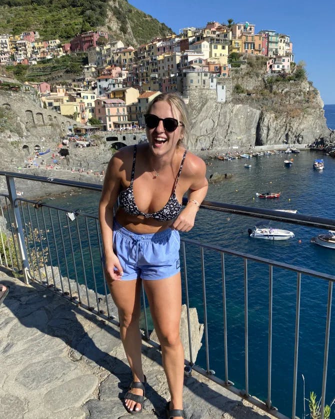 Samantha wearing a bikini top and blue shorts standing against a metal railing overlooking the blue sea and coastal region of Italy 