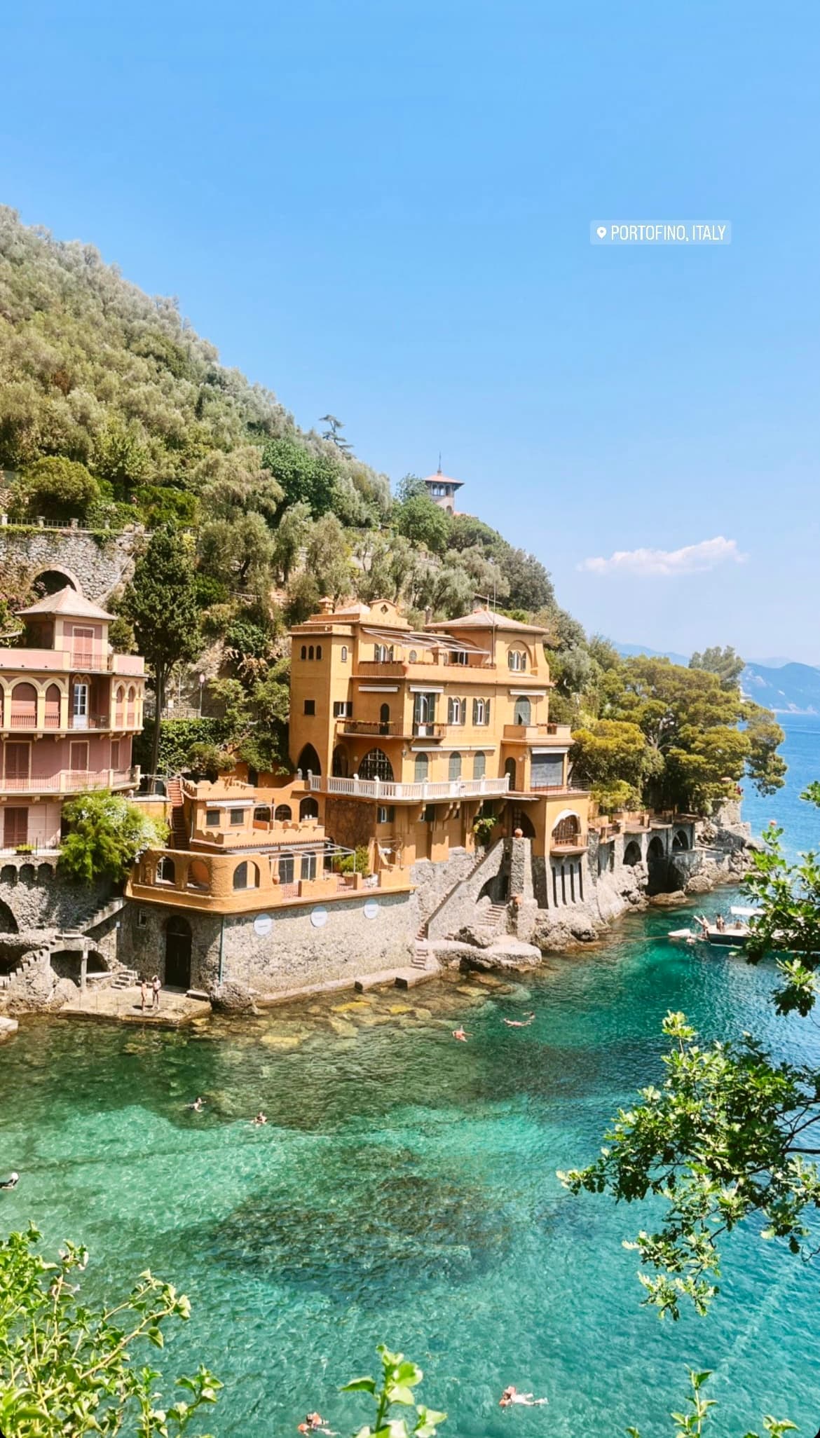 A beautiful view of an orange estate nestled into a cliffside covered in trees. There is crystal-clear turquoise waters below and trees on the right side of the image.