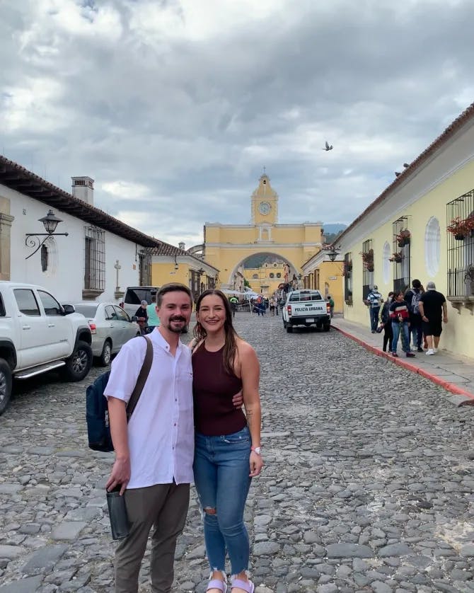 Shelby wearing a red tank top and jeans posing with a partner on a cobblestone street leading to a city center on a cloudy day