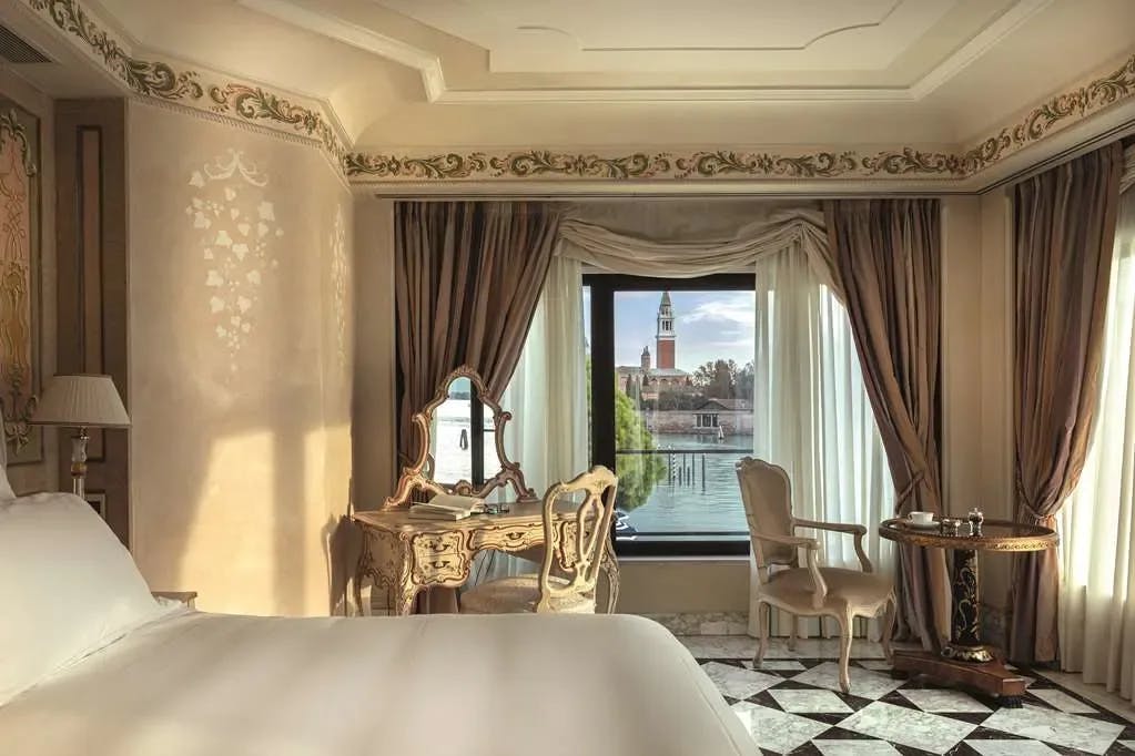 Elegant Old World furniture fills an exceptionally opulent room at Cipriani, A Belmond Hotel, Venice. Through a window, the canals of Venice and a famous church are visible