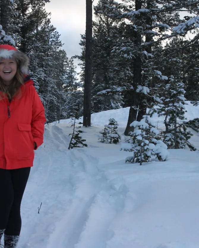 Picture of Merritt wearing a red jacket and smiling on a snowy trail surrounded by pine trees outside