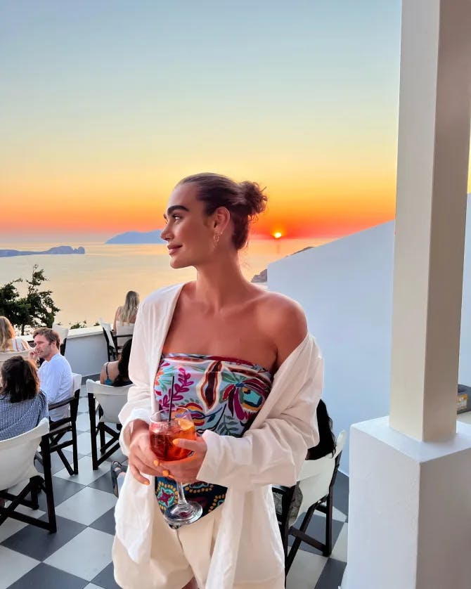 Picture of Isadora with a drink in hand standing in front of an orange sunset on a restaurant patio