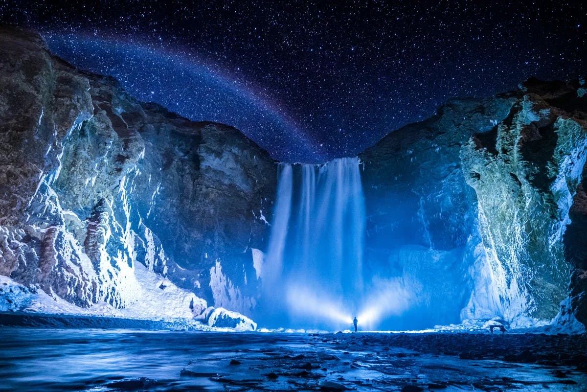 A person in front of waterfalls and the sky filled with stars during nighttime