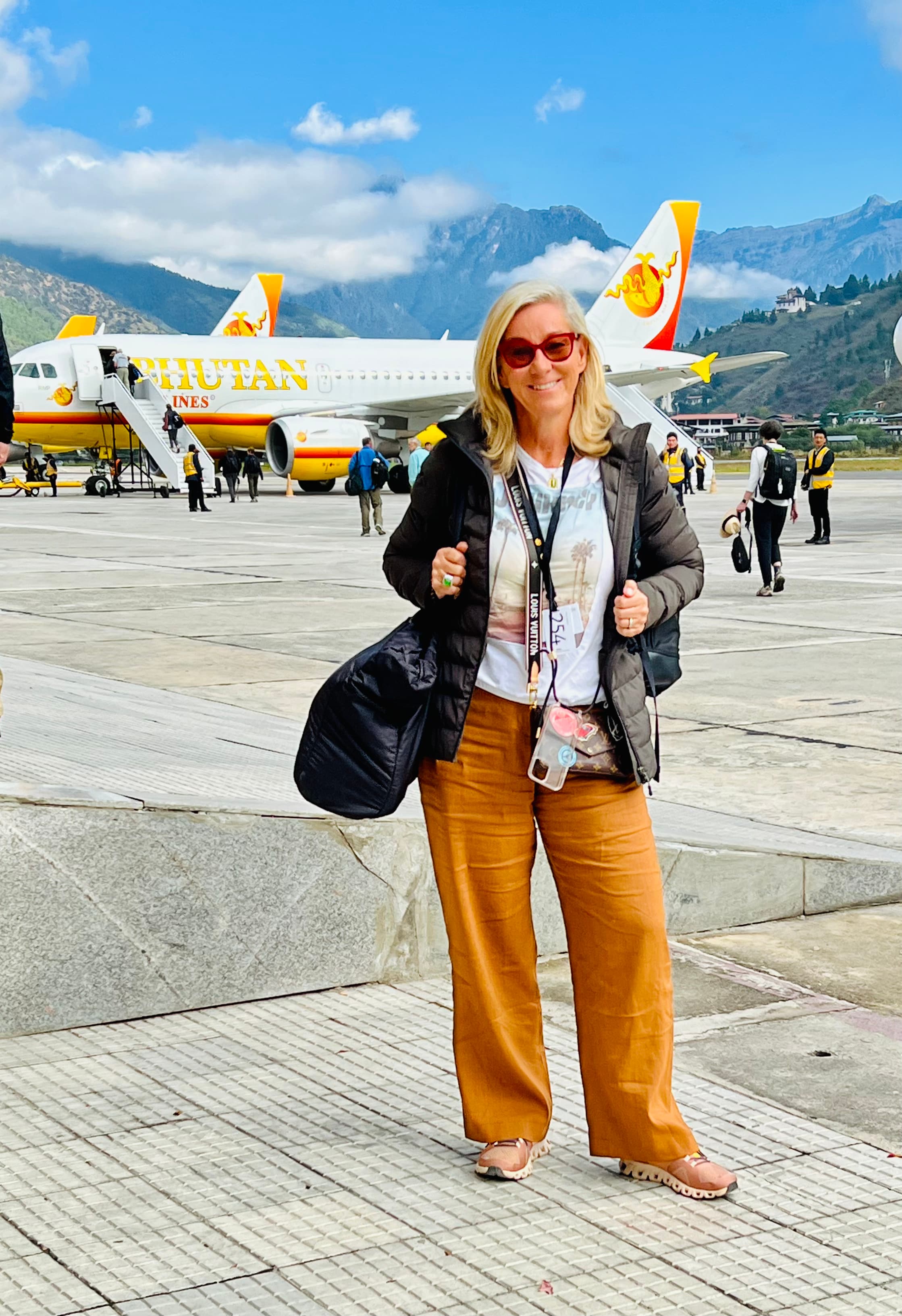 Susanne posing in front of a plane outside while wearing orange pants, a white t-shirt and jacket