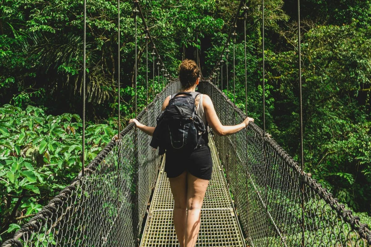 A woman in warm-weather attire and a backpack traverses a rope bridge through the jungles of La Fortuna, Costa Rica