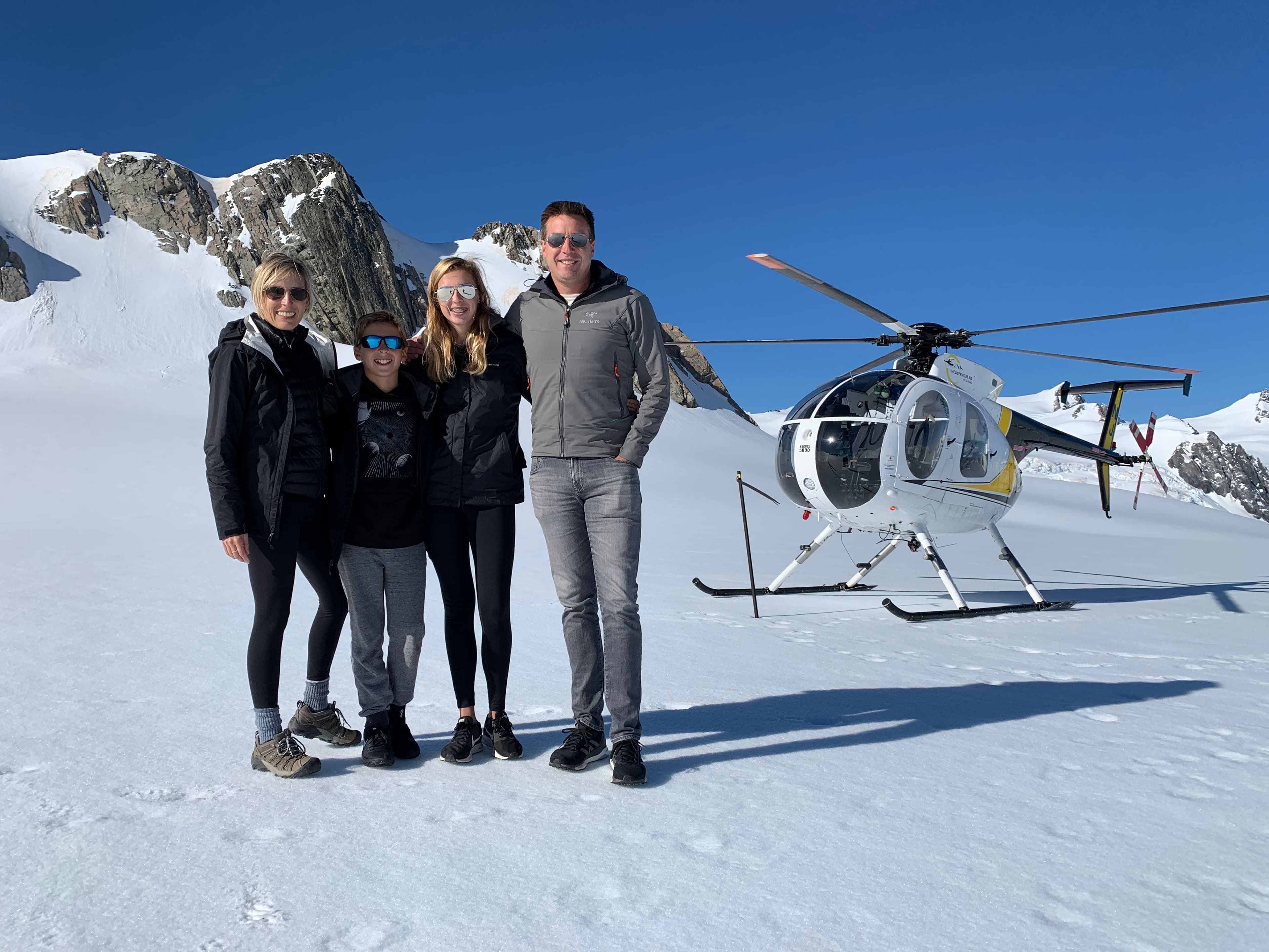 A group of people posing in front of a helicopter on top of a snow field in the mountains