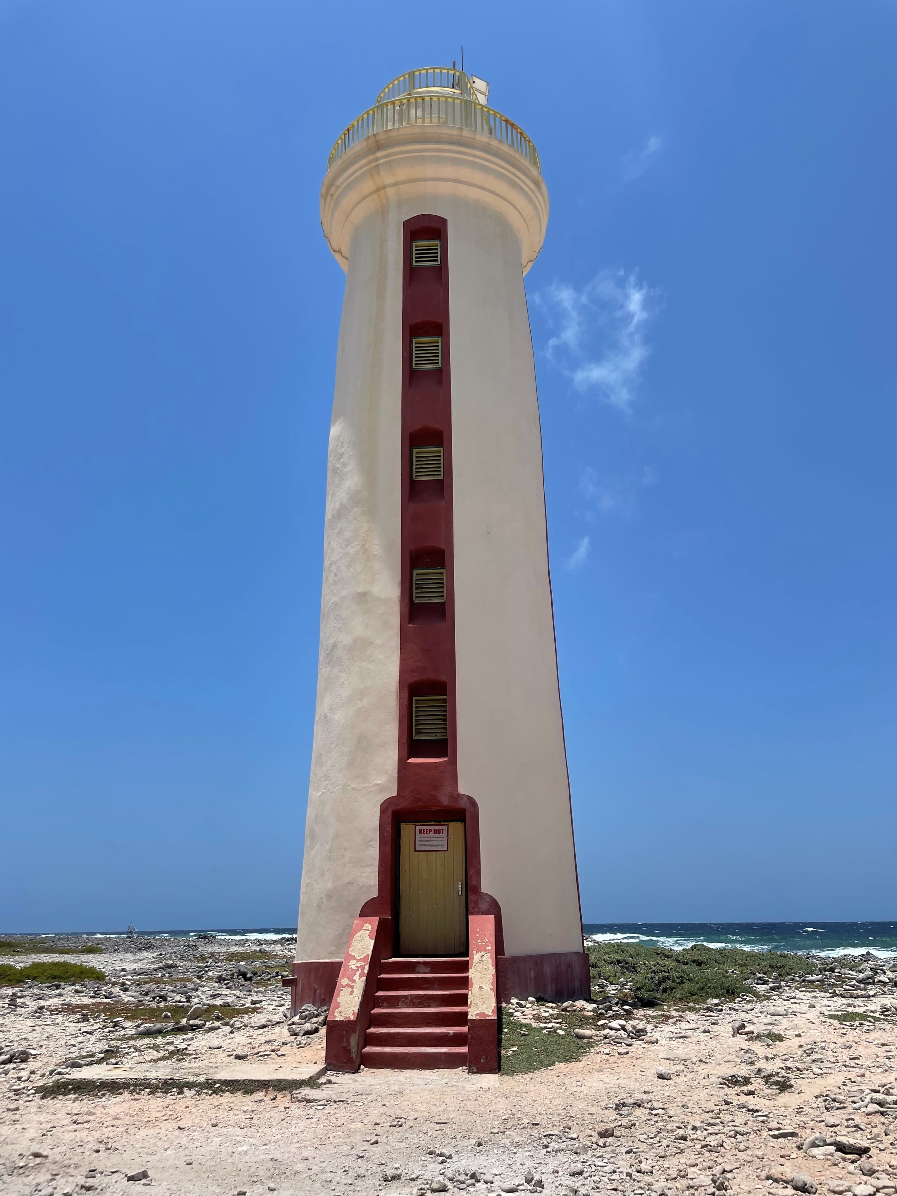 A large white tower with a red stairwell and doorframe in the middle of a desert overlooking the sea