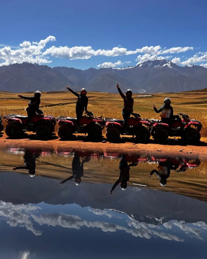 Four people pictured on bikes near a body of water reflecting the mountains and cloudy blue sky