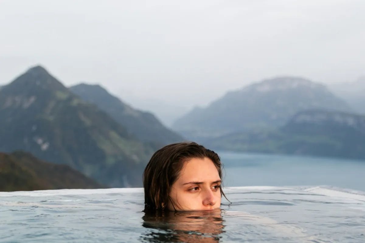A woman pokes her head out of an infinity pool. Behind her: coastal mountains rise up out of clouds for an eerie, luxe vibe