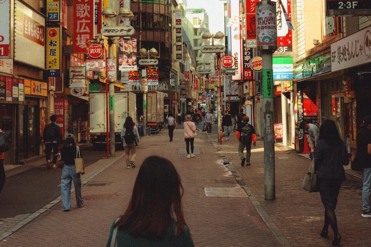 Dozens of people walk along a sign- and light-filled street in Shibuya, Tokyo while a young woman has her back to the camera