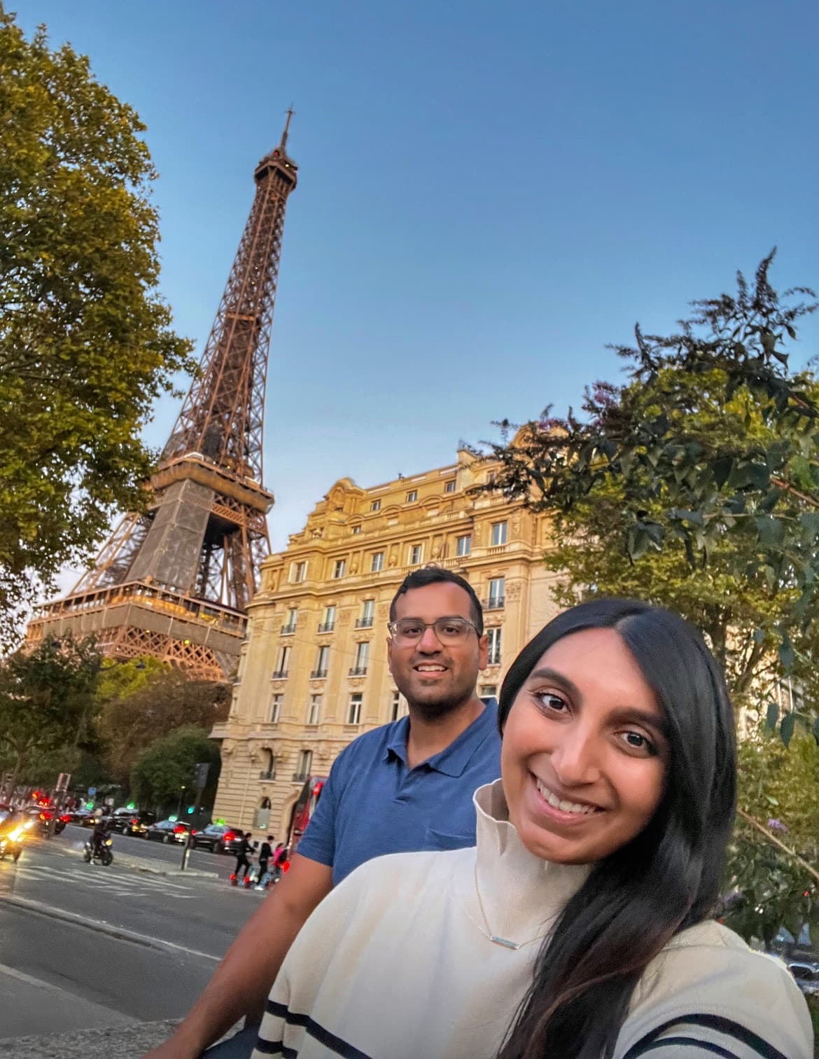 Khushbu and friend standing on a Parisian street, taking a selfie with the Eiffel Tower in the background.