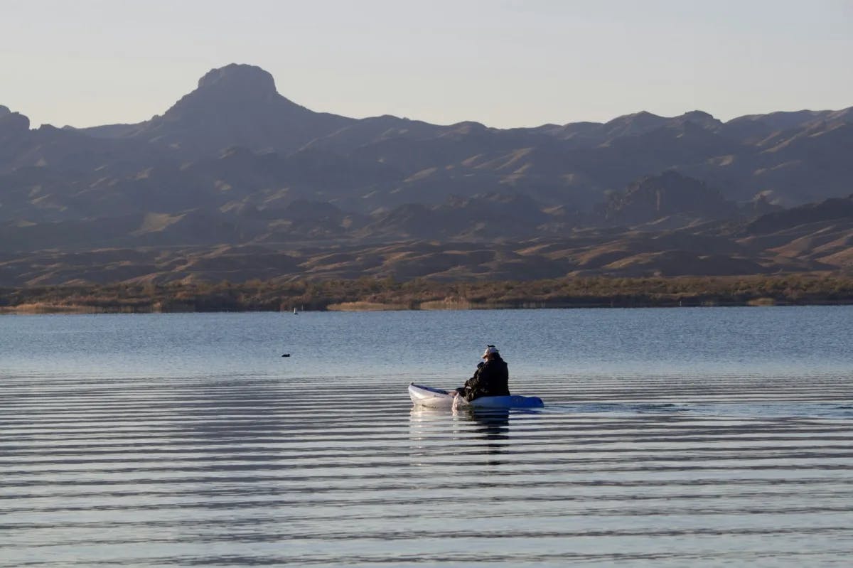 a lone kayaker paddles off the shore of Lake Havasu, with rocky hills on the far shoreline
