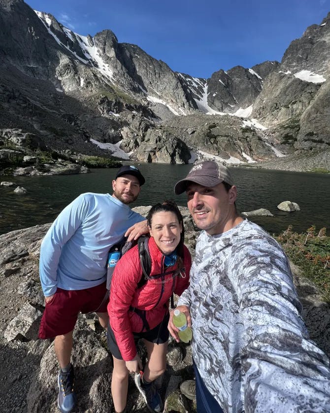 Three people posing for a selfie in front of a lake and rocky mountain dusted with patches of snow
