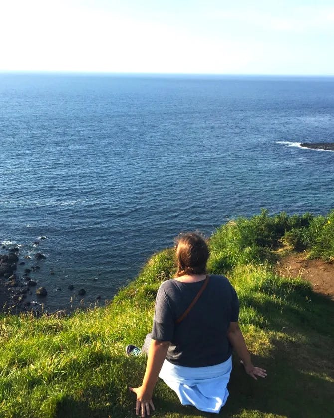 Jamie sitting on a grassy cliff looking out at the blue sea
