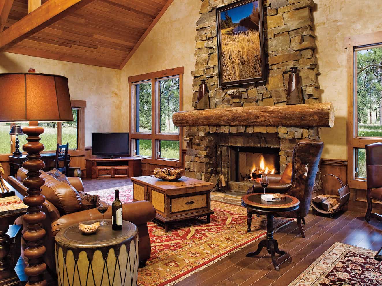 Rustic cabin living room with a stone fireplace, leather seating, wood-beamed ceiling, and large windows overlooking the forest.