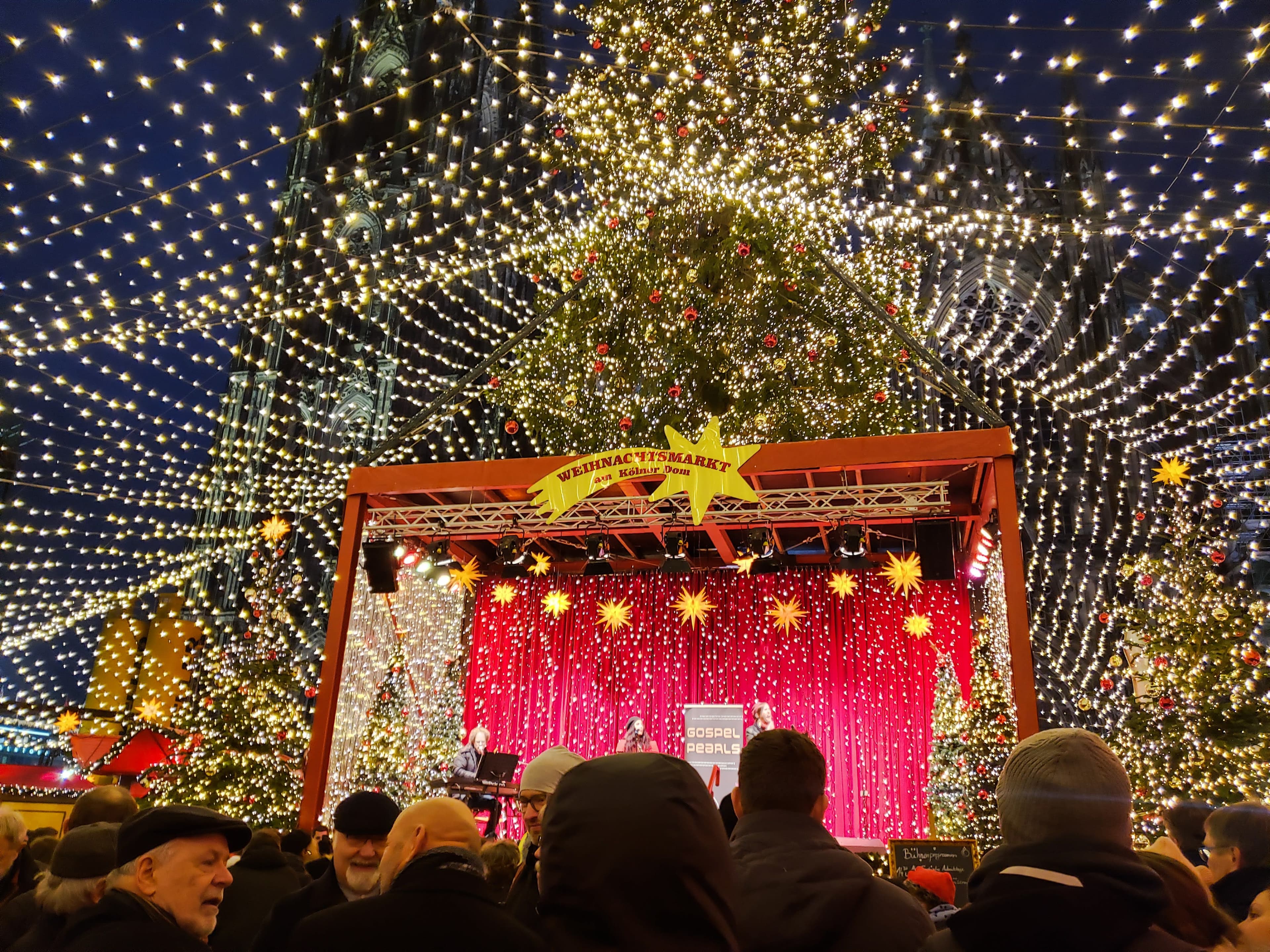 A festive scene of a decorated Christmas tree, on top of a red, pop-up stage, underneath strings of amber lights.