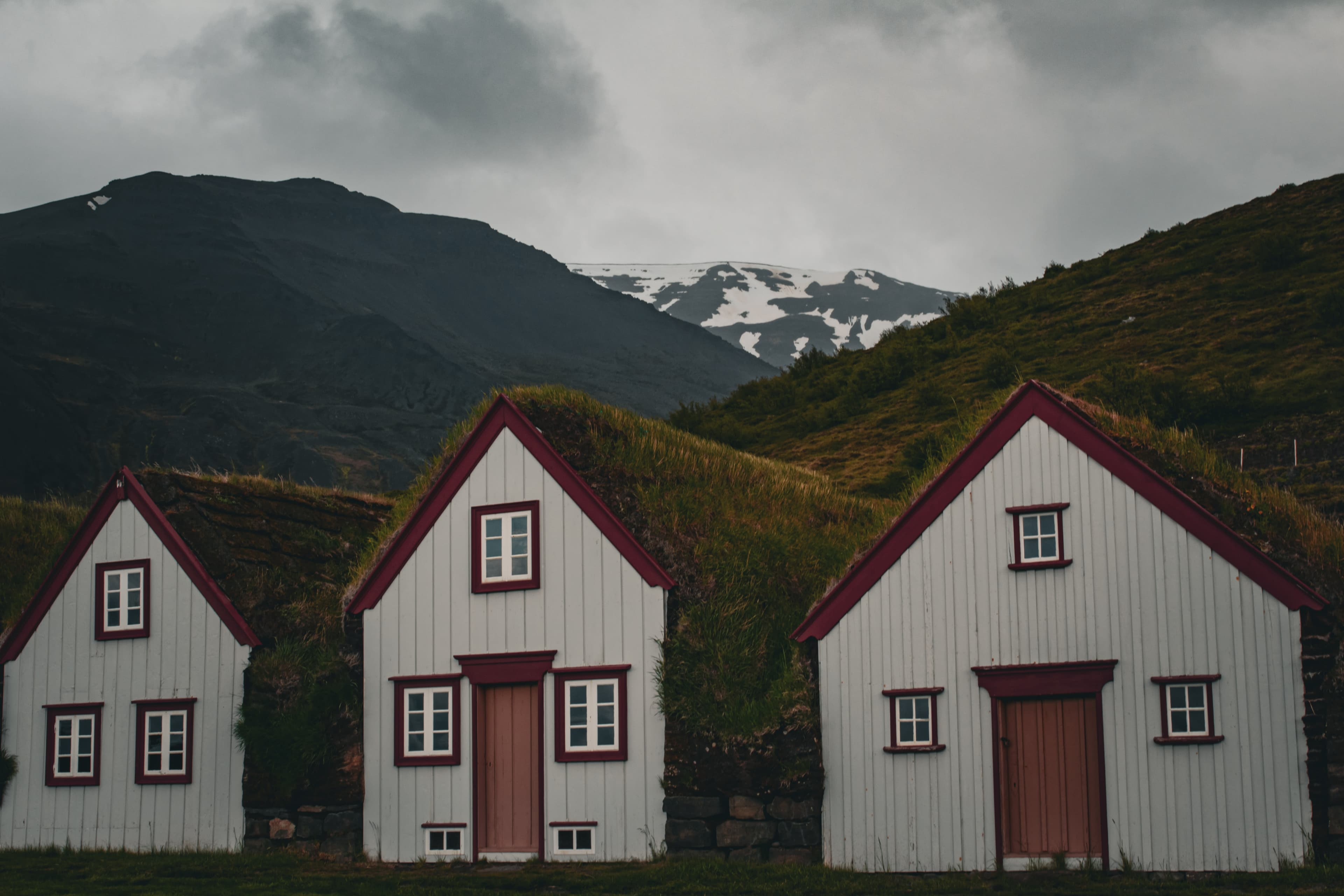 White and red cottages of Laufás Museum, with grass-covered roofs, before the mountains, on a cloudy day.