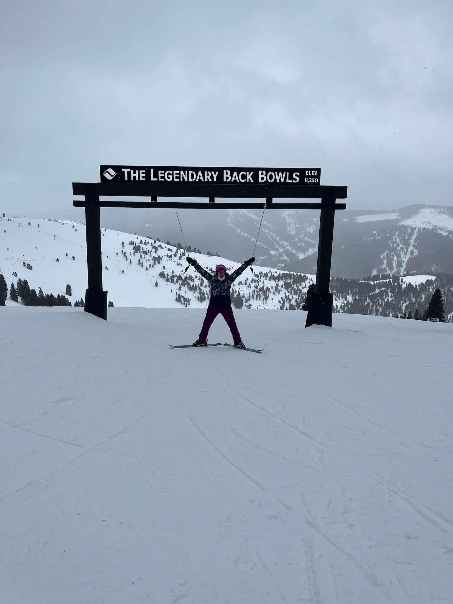 Vanessa standing underneath the wooden "Back Bowls" sign, on the slopes with her arms stretched out.