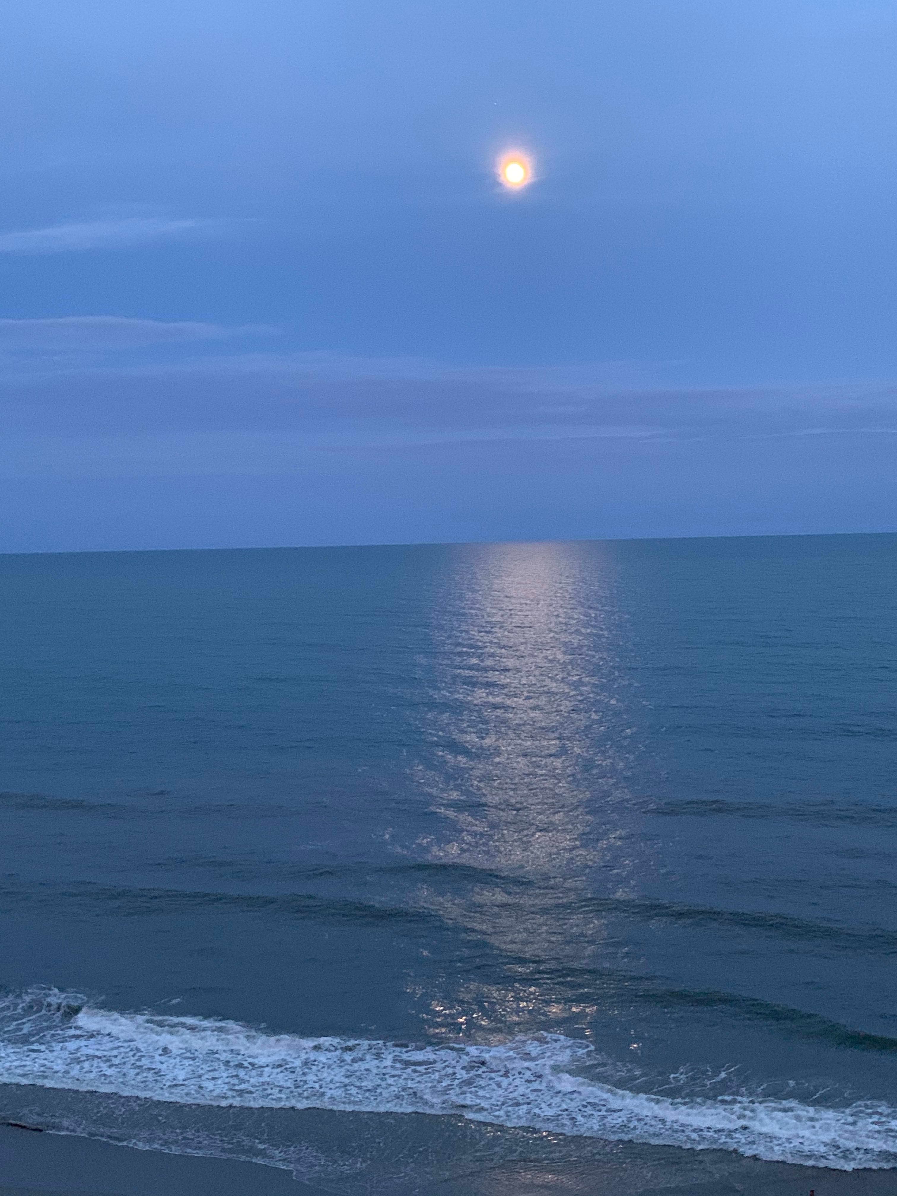 A scene of the amber moon peaking through the clouds, and reflecting off of the sea at dusk.