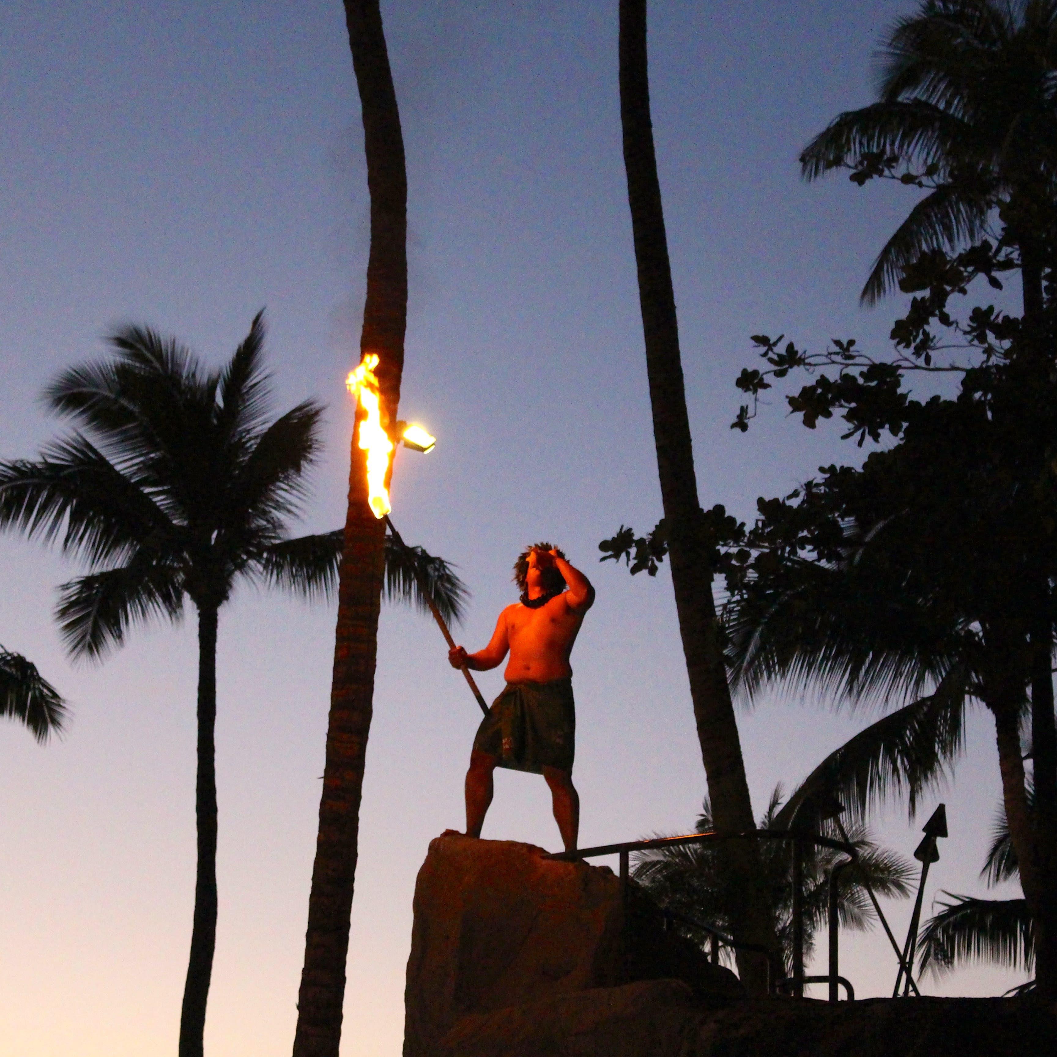 A man in traditional dress standing ona rock under palm trees at night doing a fire show