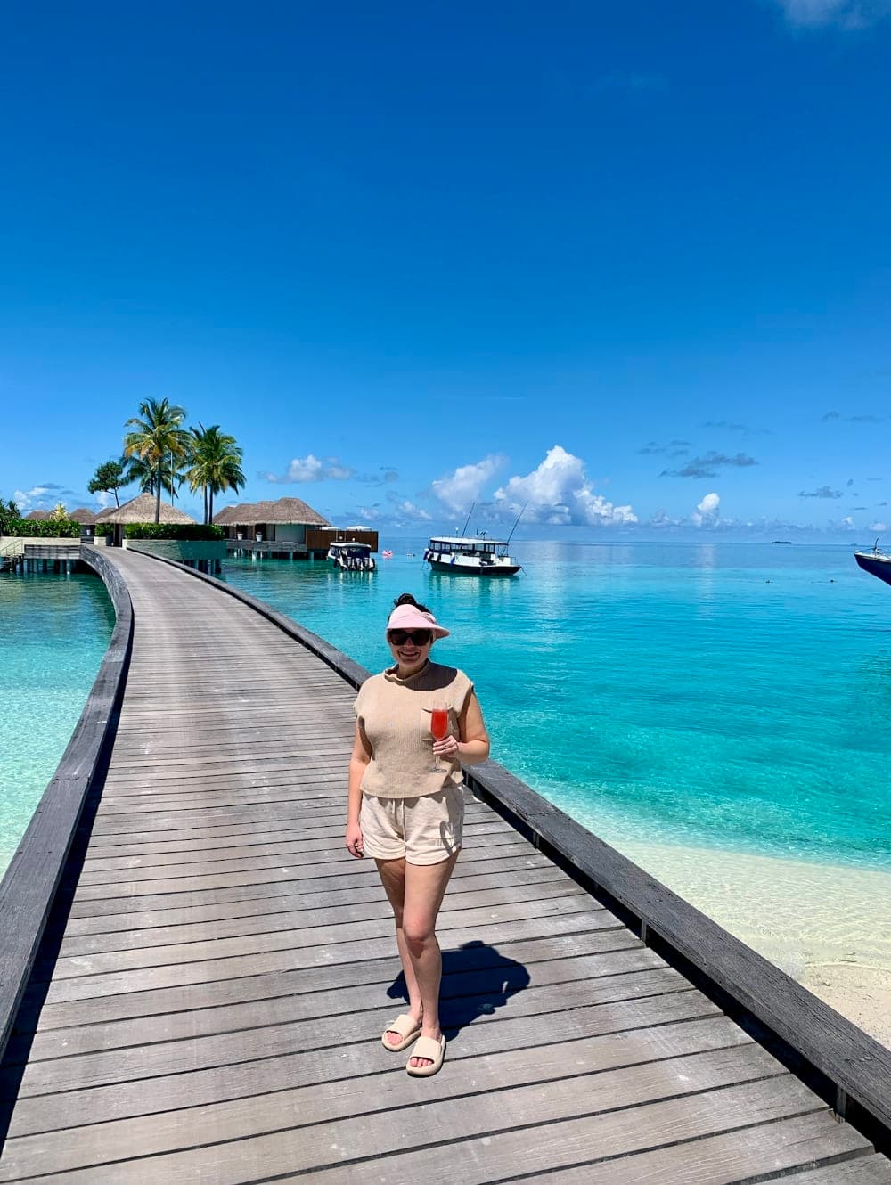 A woman posing on a wooden path with a drink in hand and view of palm trees, bungalows and the turquoise waters in the background.
