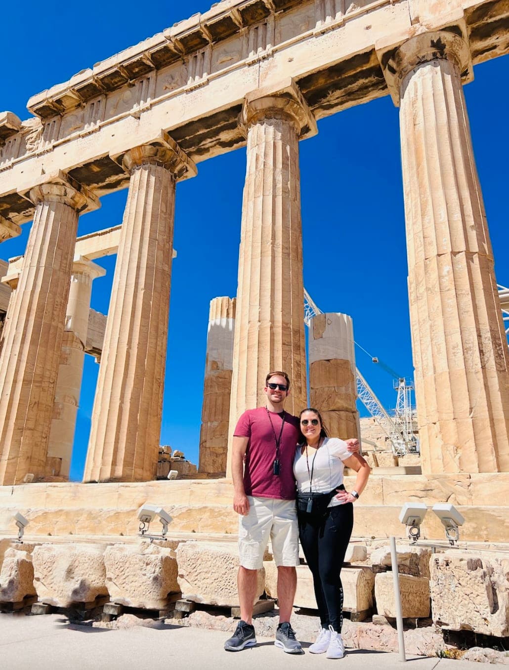 A couple posing together in front of the stone pillars of the parthenon with a blue sky in the background.