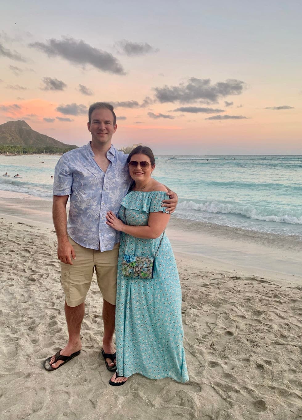 A couple posing together on a beach with the ocean, a volcano and a light pink sunset in the background. She is wearing a long blue dress and he is wearing a blue top and khaki shorts.