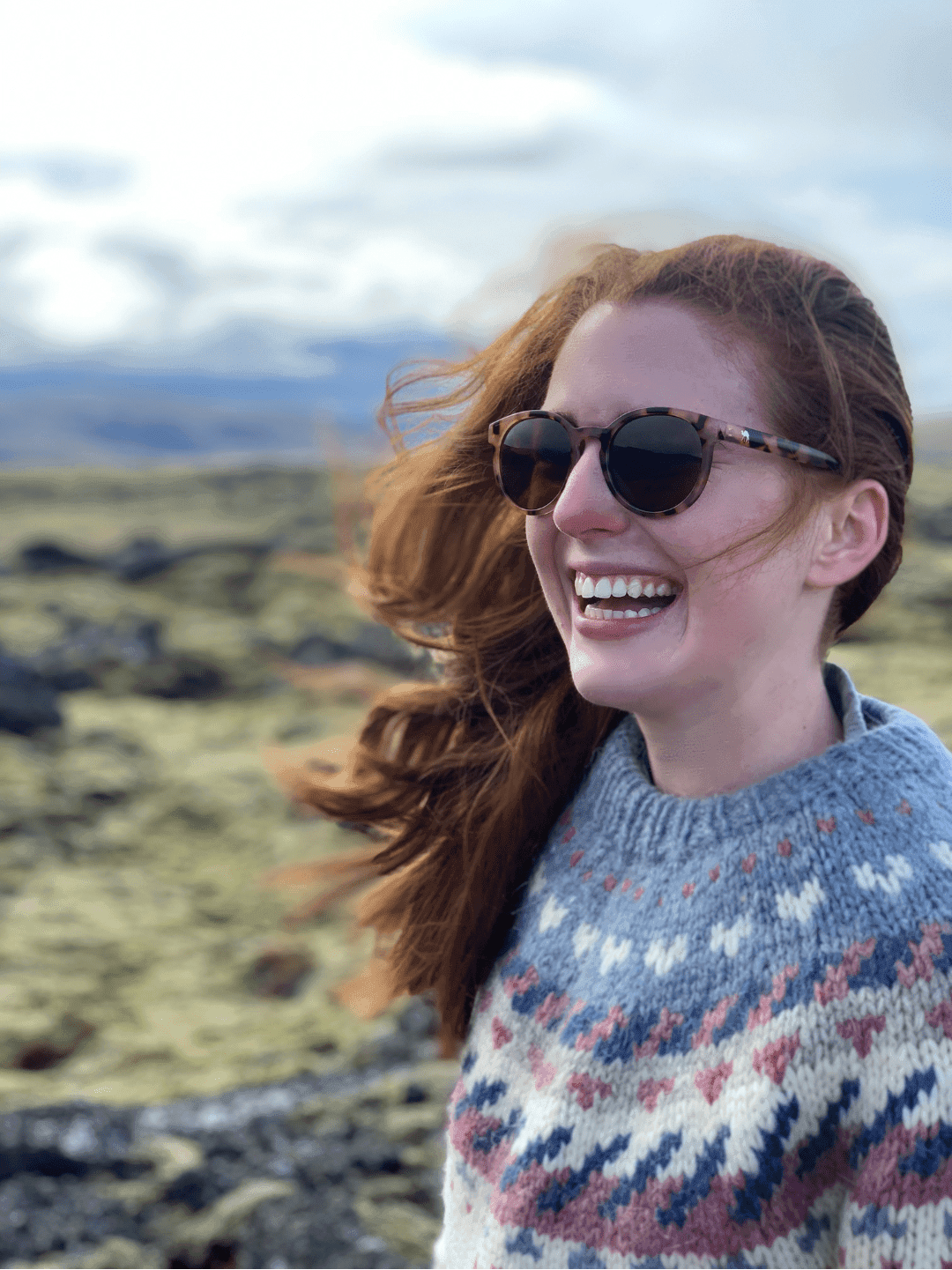 A portrait of Hannah in a sweater and sunglasses with a green hilly background in Iceland
