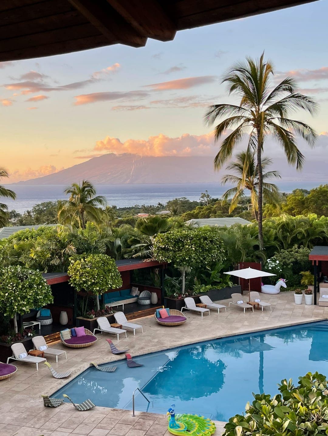 A pool with poolside chairs and a background of green trees and the ocean in Hawaii