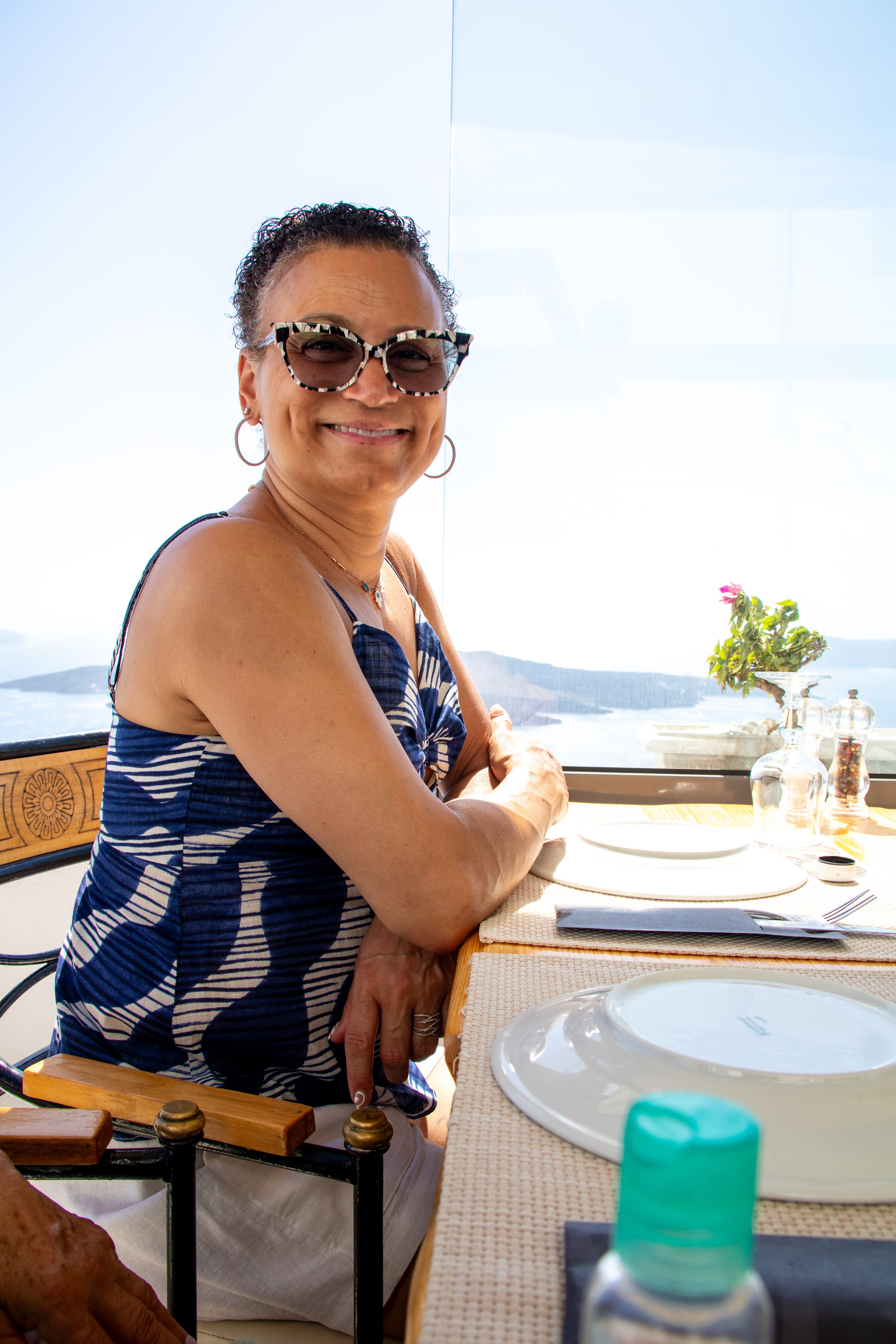 Adrianna in a blue tank top and sunglasses sitting at a table on a restaurant patio next to the sea