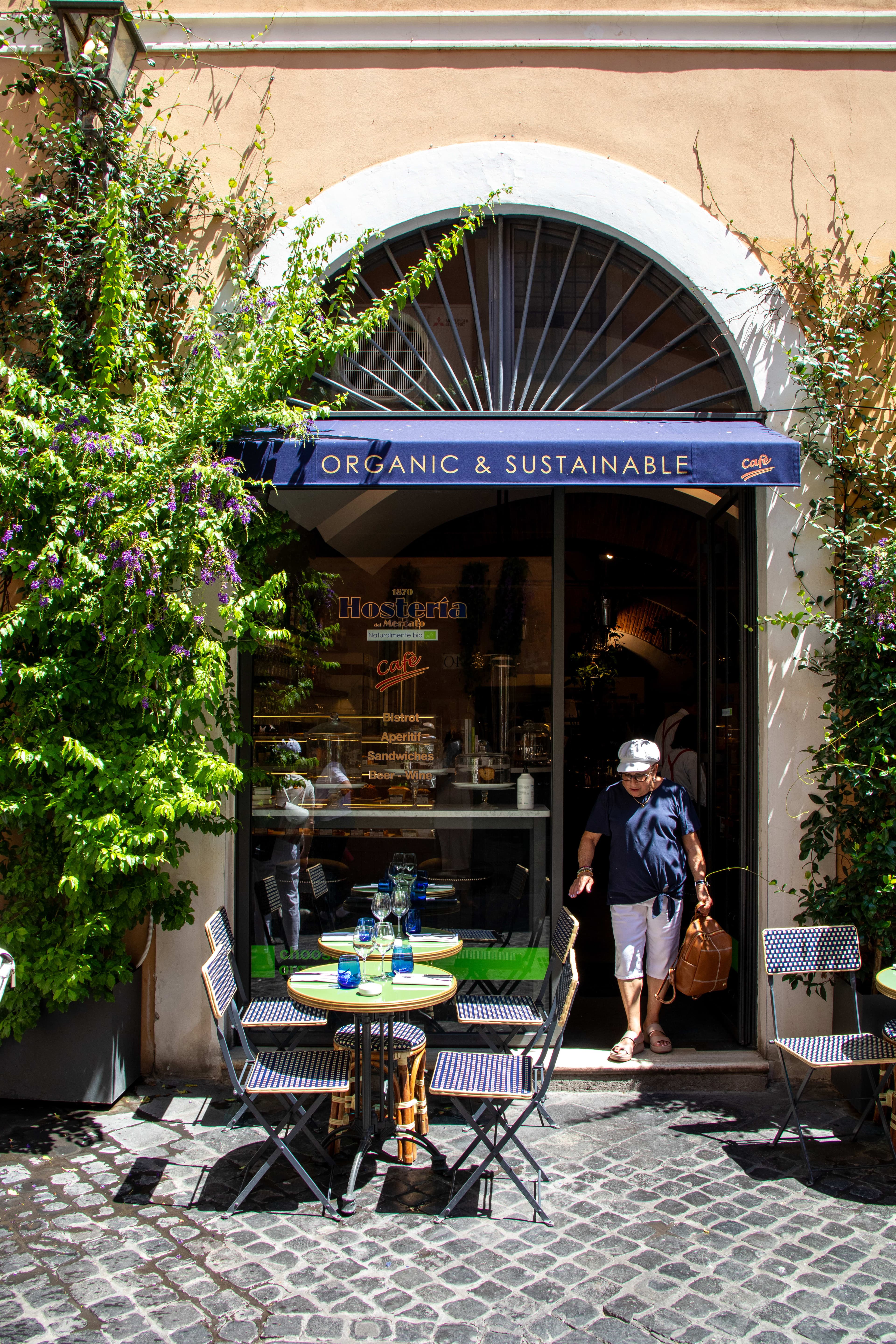 The exterior of a restaurant with a table and chairs on the patio and a blue awning that says "organic & sustainable"