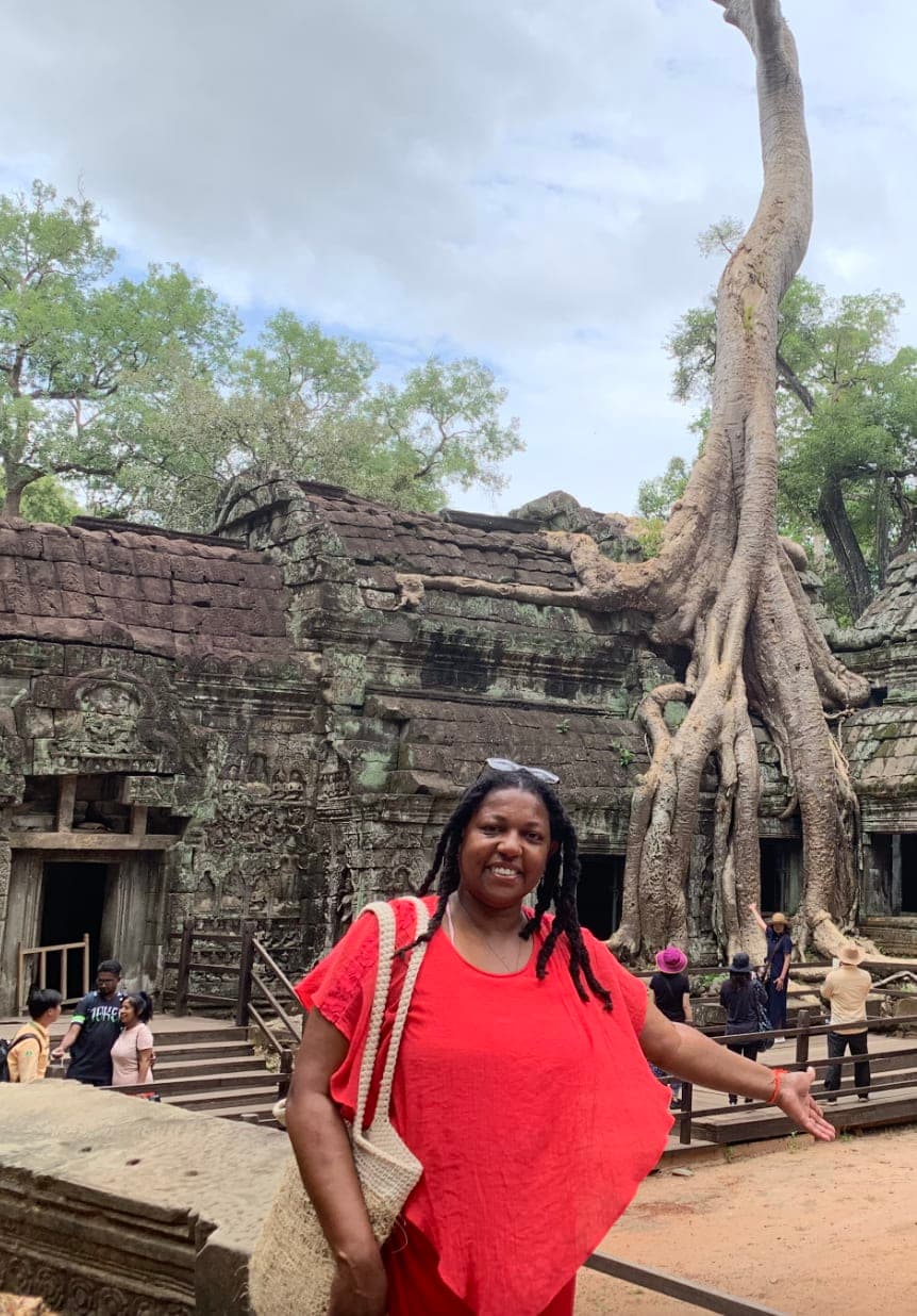 Lynore posing in a red top outside in front of an old stone temple and trees with left arm outstretched.