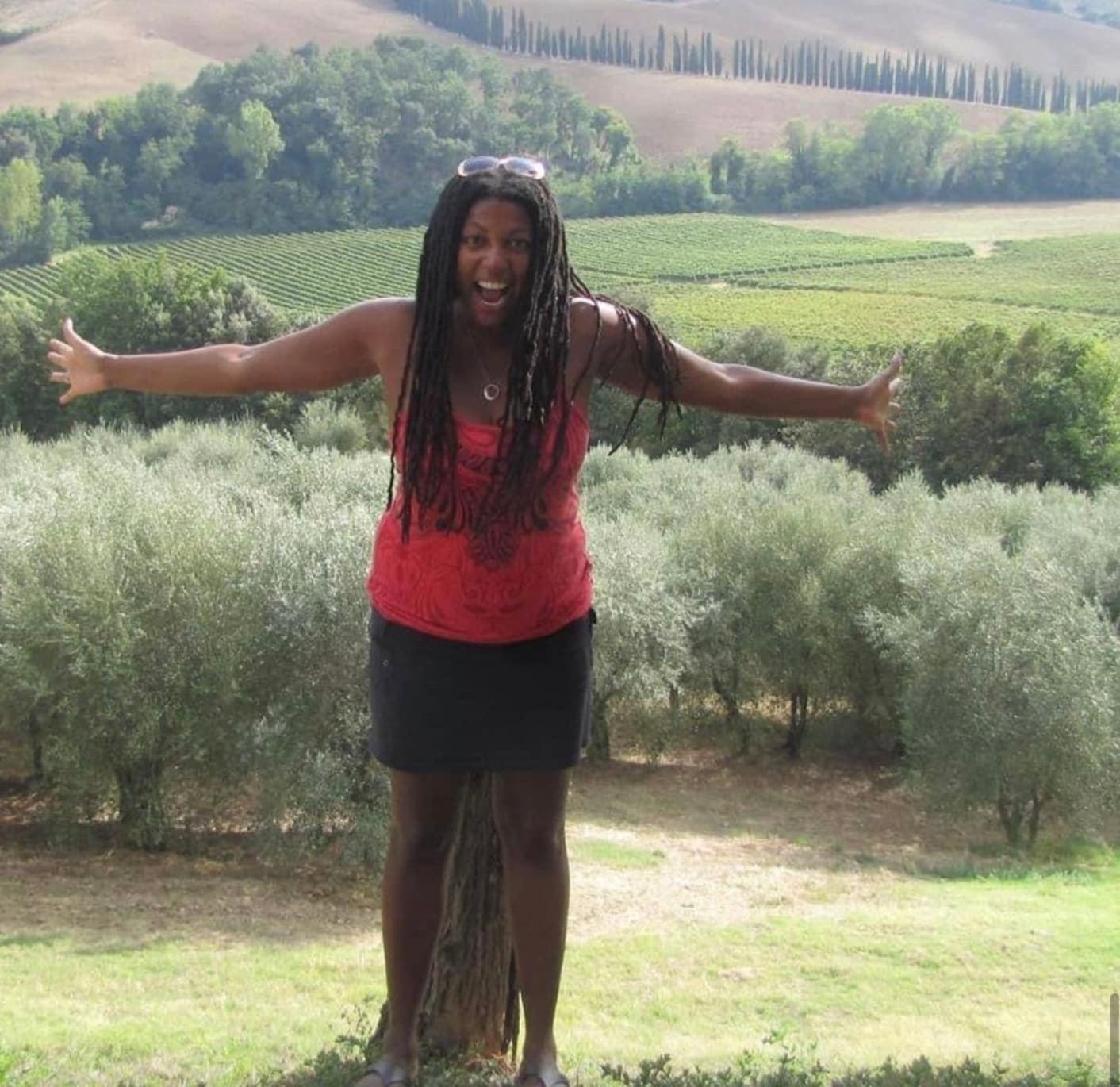 Lynore standing outside in front of rolling hills and beautiful green trees with her arms outstretched.