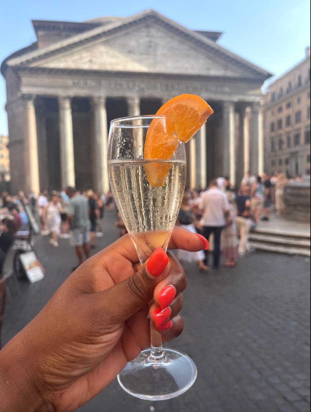 A person with pink nail polish on holding a champagne flute with an orange slice on it. There are people walking around a historic square in the background.