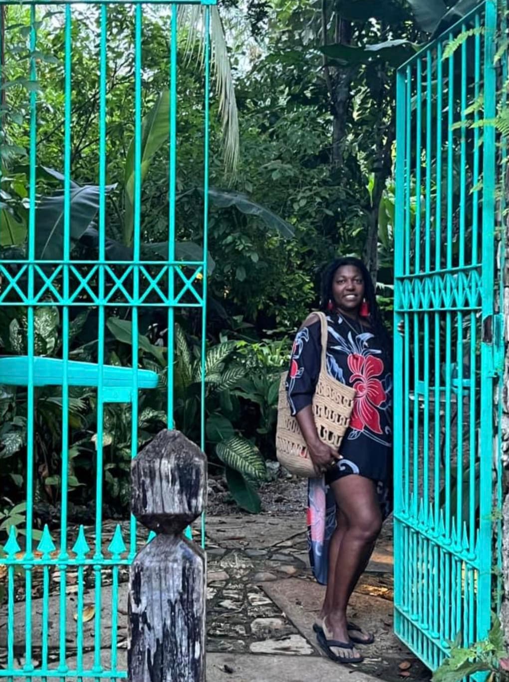 Lynore posing in front of a turquoise gate and trees while wearing colorful clothing.