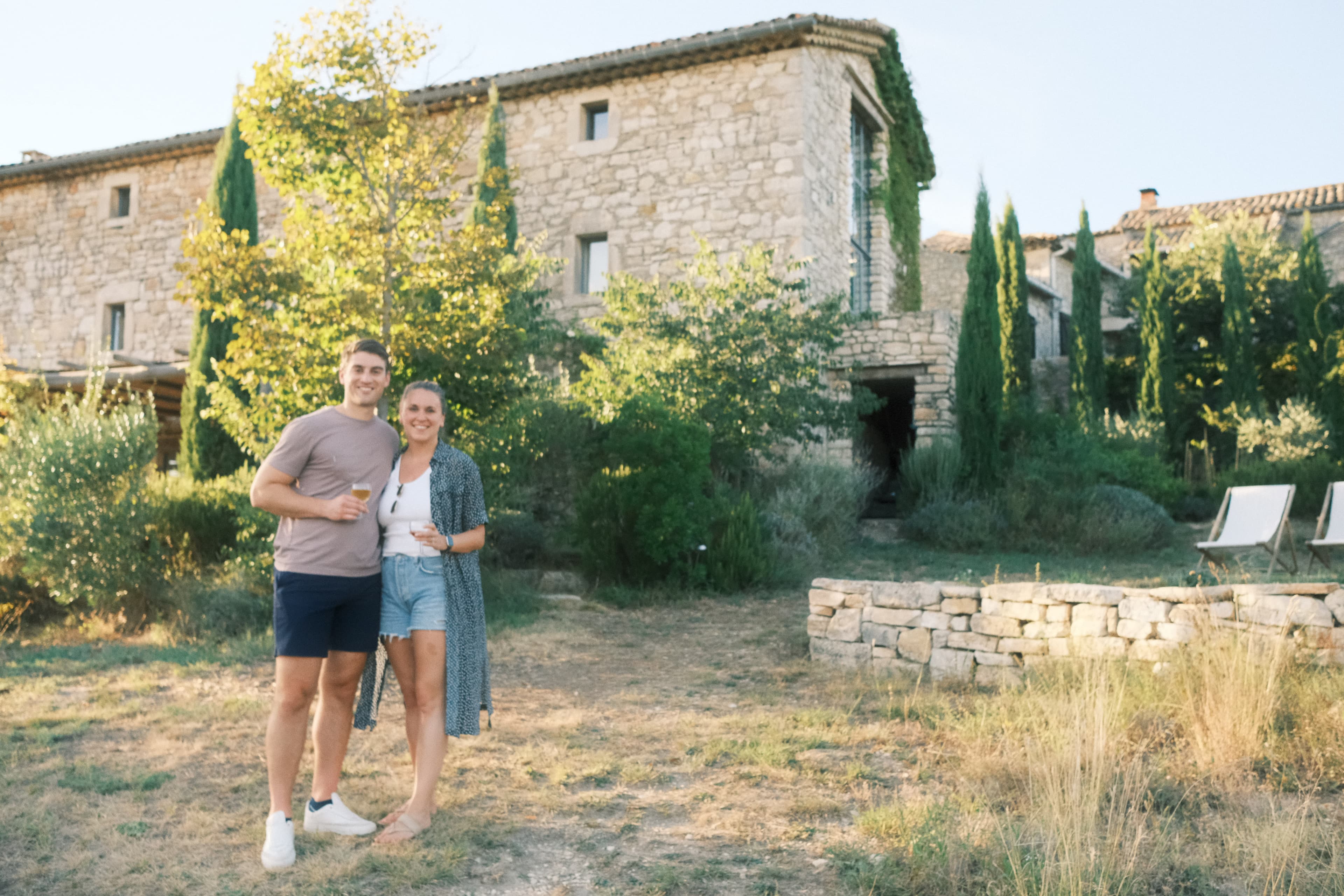 Gloria in shorts next to a man holding drinks in front of a stone building