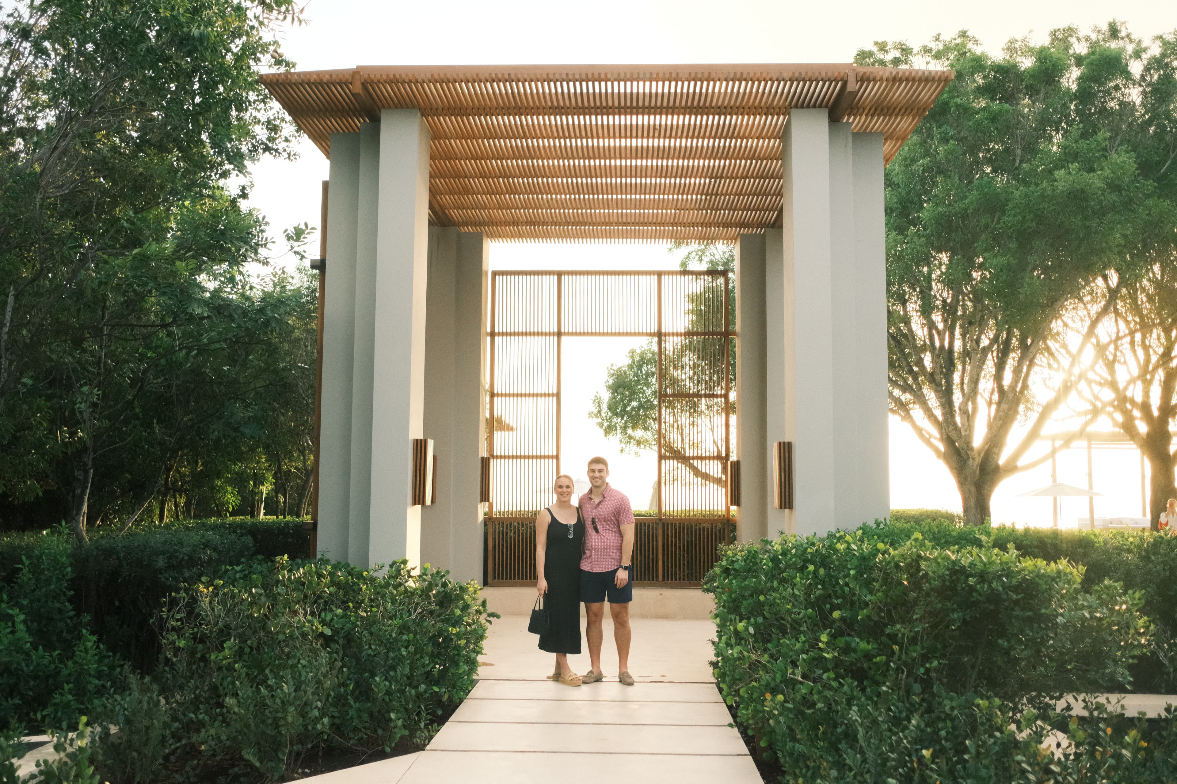 Gloria in a black dress standing next to a man in a pink shirt in front of a large stone gate with a thatched awning on a path between bushes