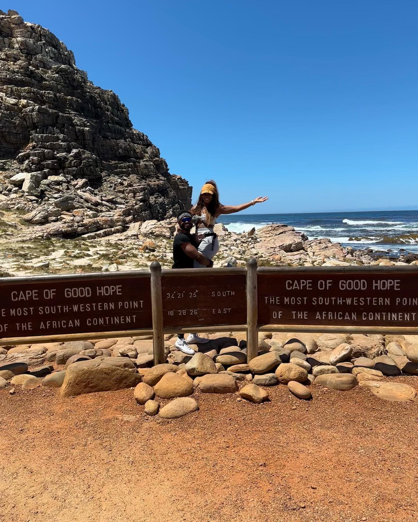 A man holding up Sitaara behind the Cape of Good Hope sign with rocks and the ocean in the background