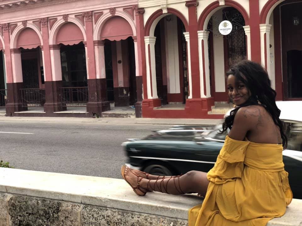 Sitaara in a yellow dress sitting on a stone wall next to a street with red archways