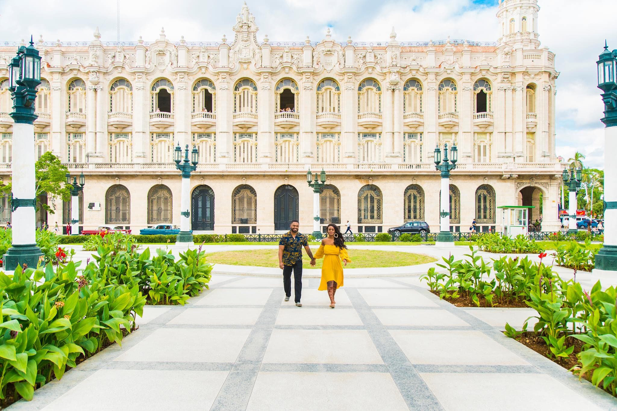 Sitaara in a yellow dress walking hand-in-hand with a man down a path in front of the Grand Theater of Havana, a large ornate building