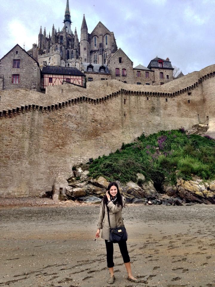 Carissa in a green jacket standing barefoot on a beach in front of a city wall with a castle behind it.