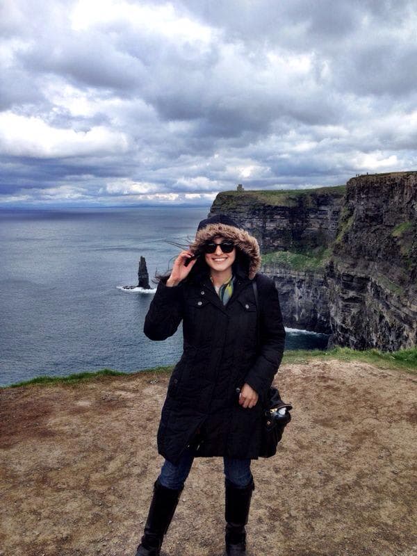 Carissa in a black jacket with a fur hood standing on a cliff in Ireland with the sea behind her.