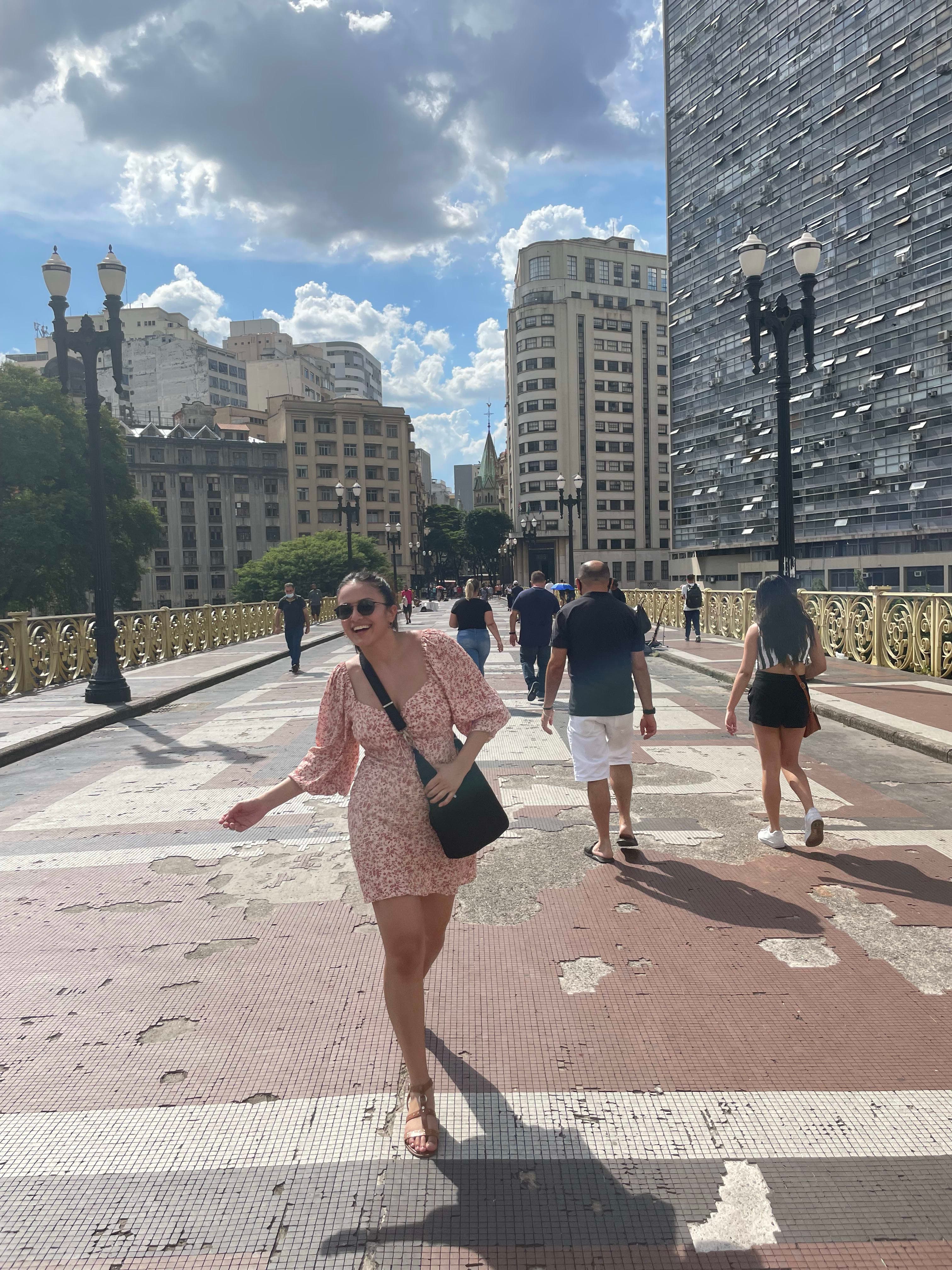Carissa in a pink dress walking across a bridge with a city behind her.