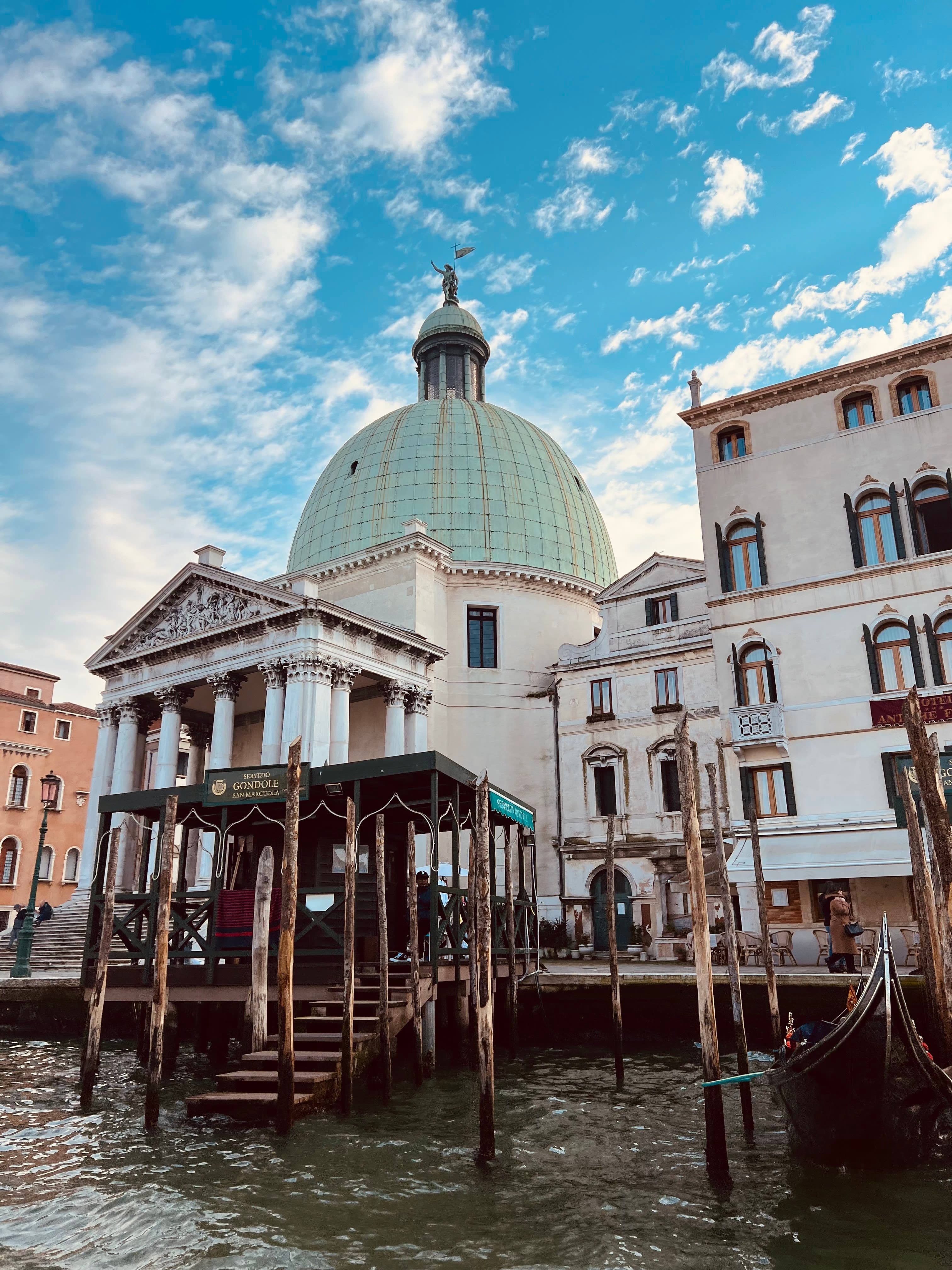 A scene in a city from the river with a small wooden pier and a white building behind with a green dome