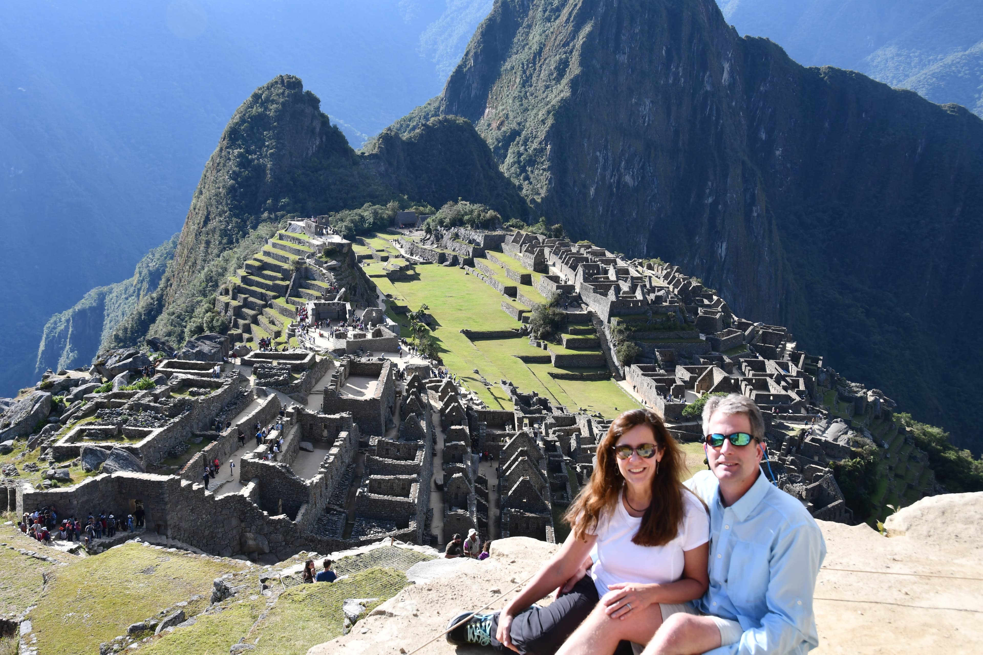 A couple posing in front of Macchu Pichu during the daytime. It's bright, green and rocky. They're both wearing sunglasses and cool toned clothing.