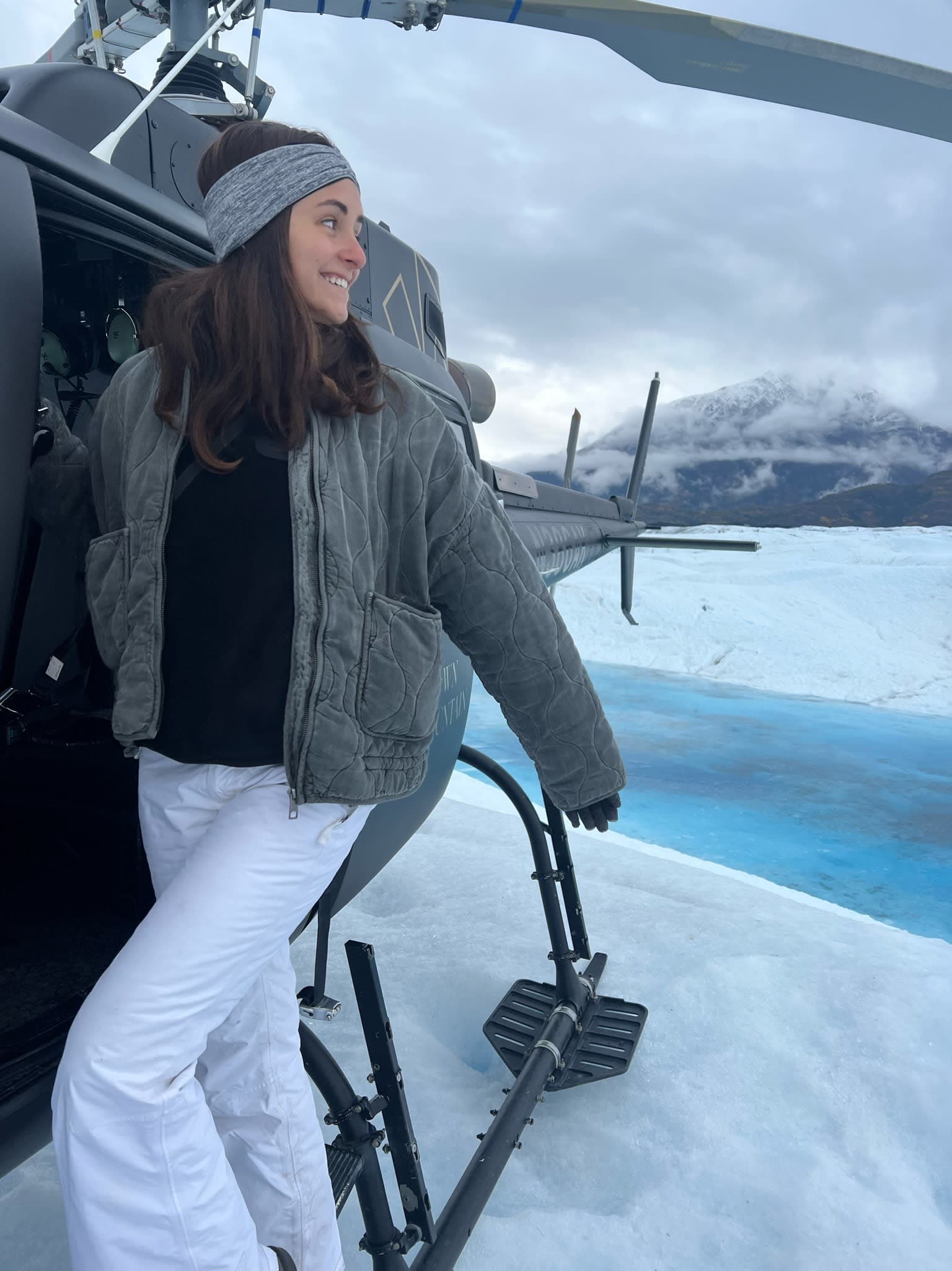 A girl in winter clothes posing out the side of a parked helicopter on top of a glacier.