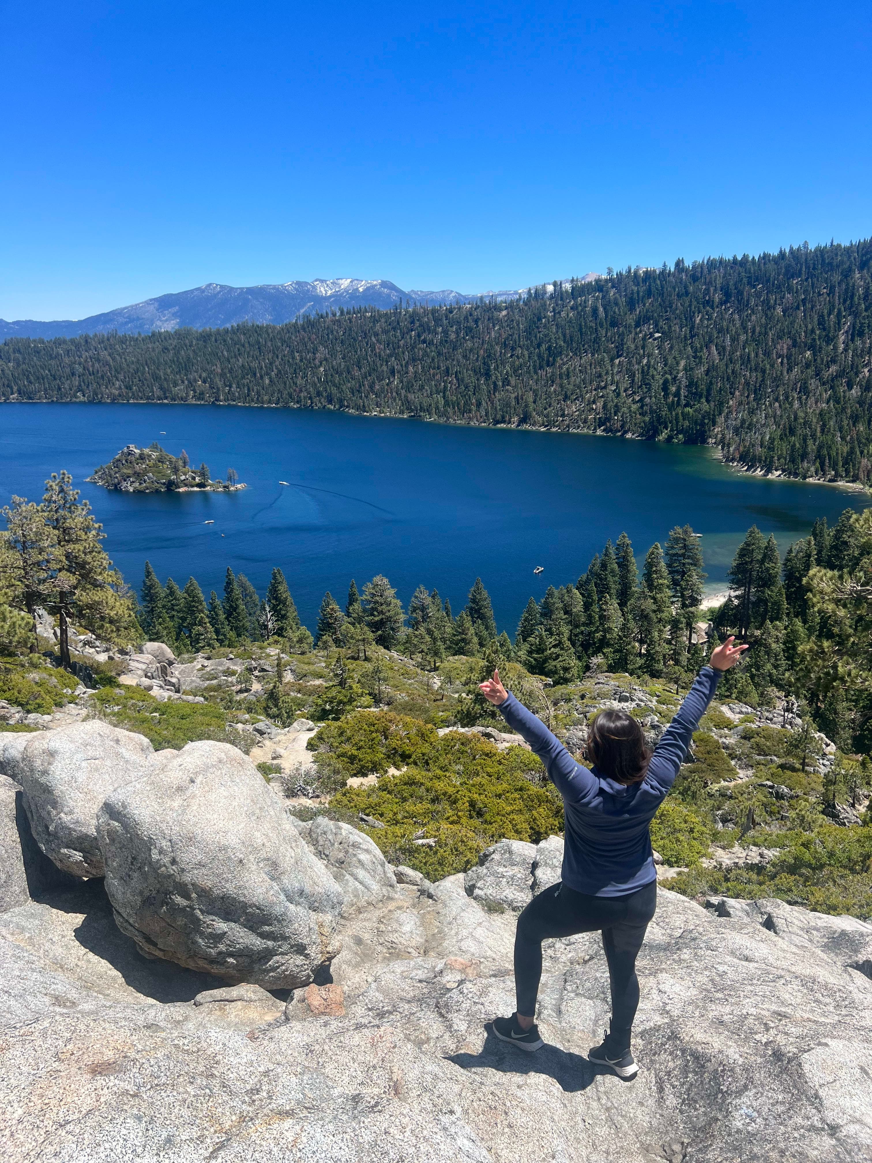 A picturesque view of travel advisor Yiselle from behind standing with her arms in the air on a rock looking down at a lake surrounded by lush green landscape