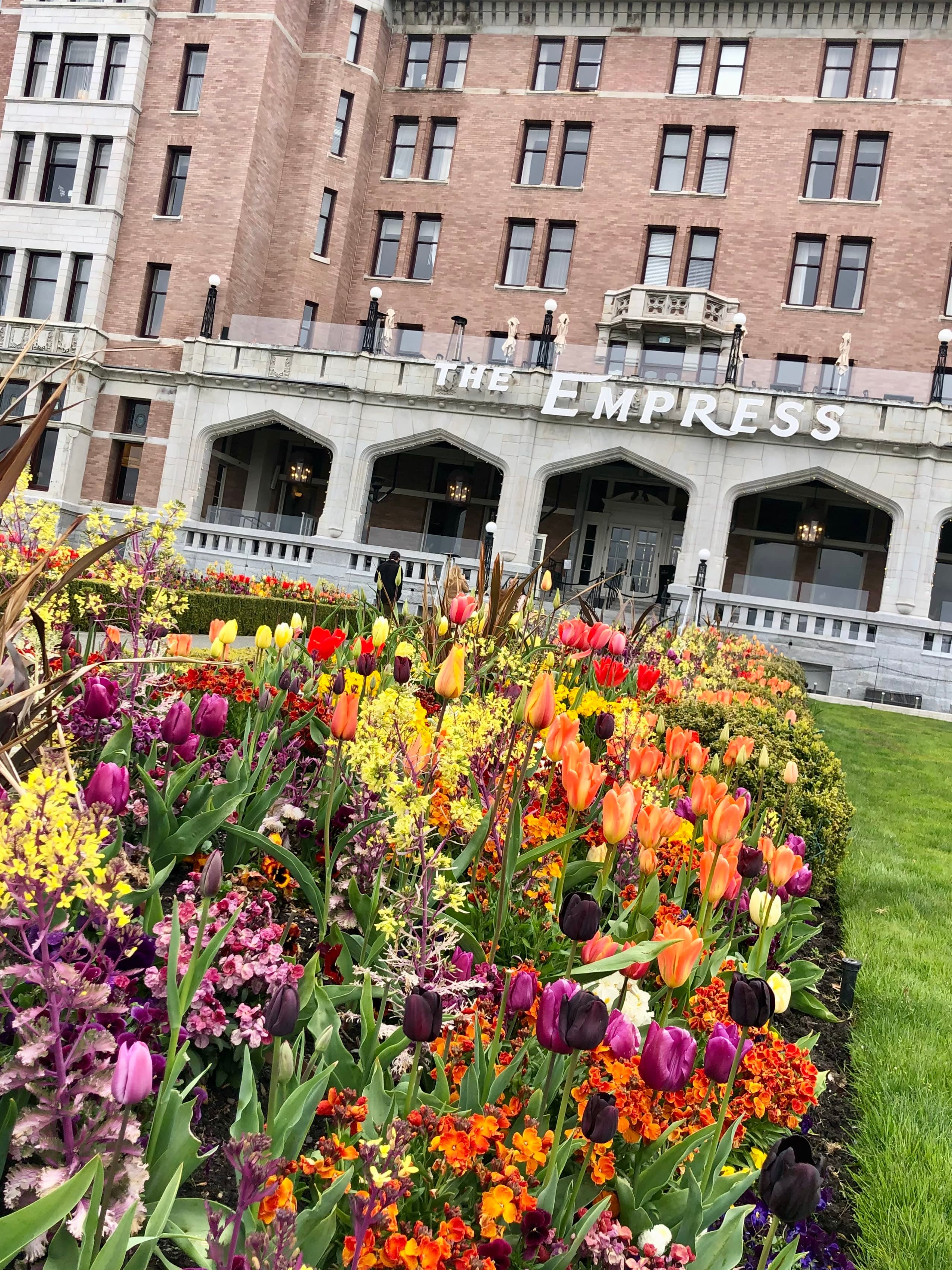 A bed of colorful flowers on a grassy square in front of a red and white building with The Empress sign.