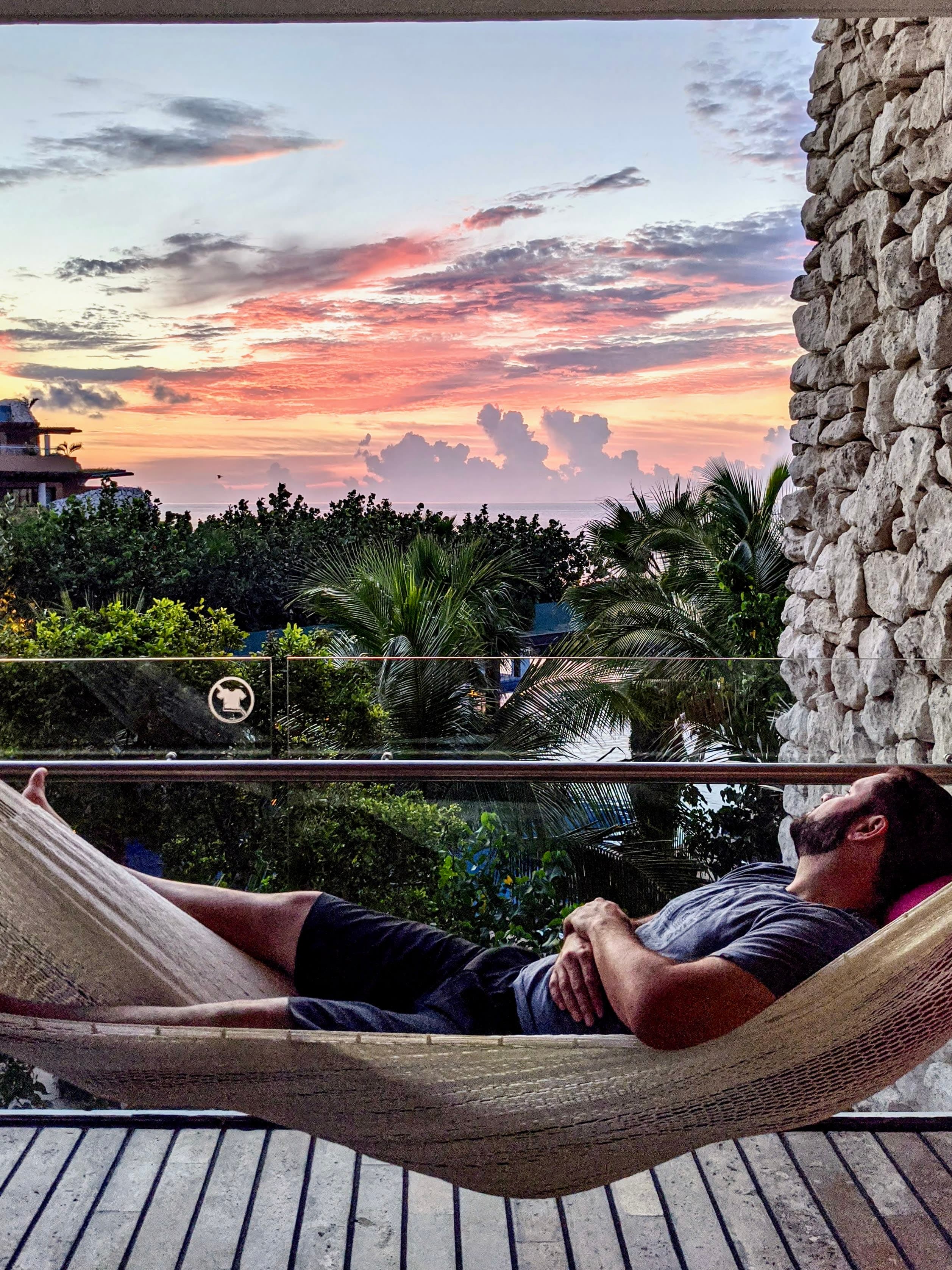 A man lying on a hammock on a balcony with wooden deck and stone wall overlooking a beautiful sunset view